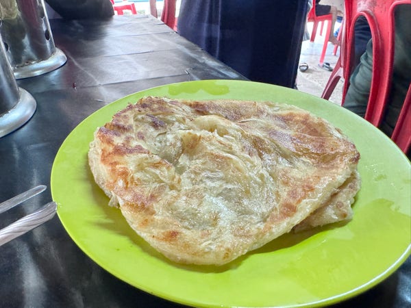 A roti canai on a green plate on a restaurant table, with utensils and teapots in the background.