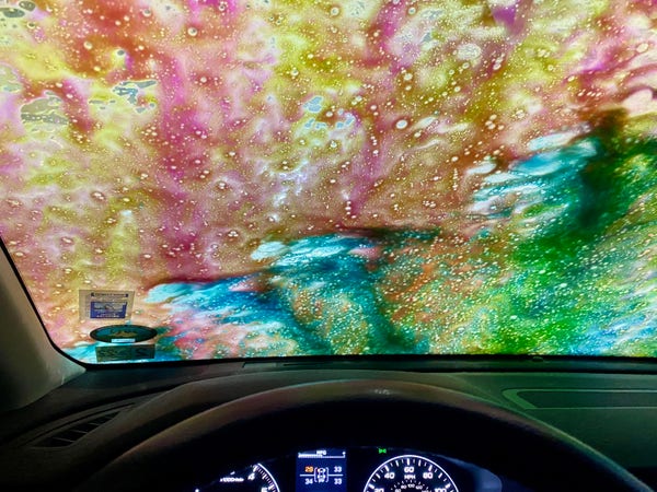 view from behind the drivers seat, the car is parked in a car wash and multicolored soap runs down the windshield