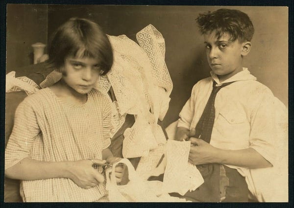  The image appears to be an old photograph, likely taken in New York, as indicated by the text at the bottom. It shows two children in front of a backdrop that resembles a wall with a doorway or window frame. The child on the left is wearing a dress and has a somewhat displeased expression. The child on the right is dressed in a suit and tie, holding a piece of paper or fabric in their hands, which could be a prop or part of an investigation. There's no visible text within the image itself to provide more context about its content or purpose. The style of the photo suggests it may be from the early 20th century based on the clothing and the quality of the image.