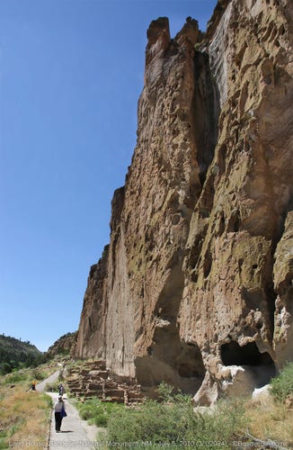 An asphalt footpath, bottom left, parallels a tall, sheer cliff of soft volcanic rock that is pierced by natural and human made holes; at the foot of the cliff are stone ruins of an ancient puebloan long house. Clear blue sky.
©BosqueBill.com