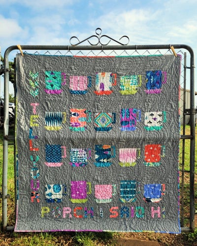 A small quilt hangs on a garden gate - the day is sunny. The quilt is made up of multi-colored fabrics in pinks, aquas, corals, navy blues and greens on a solid gray background. It has a pattern of mismatched cups and saucers, bordered on two sides by quilted text that reads: Tell Your Porch I Said Hi.