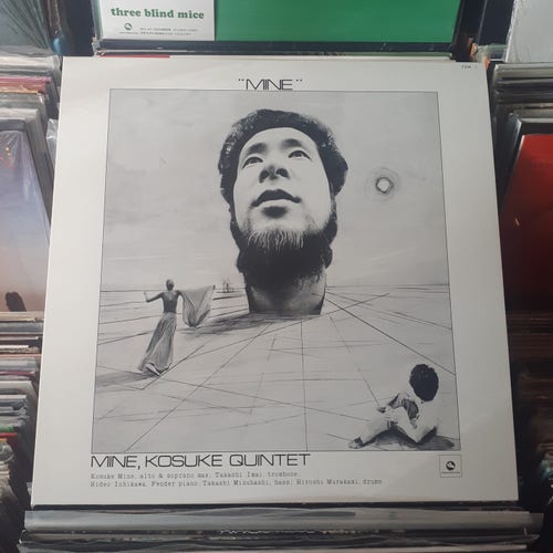 Album cover features a photo of KM's giant head protruding out of a surreal landscape. In the foreground, there's a woman with a robe or something on her extended arm walking toward the giant head. There's also a guy sitting in the bottom right corner, looking at the giant head.  It's all a bit difficult to describe, but that's the gist of it.