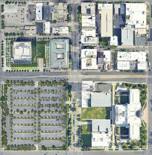 A screenshot of Google aerial imagery in an urban area. There are 4 blocks. The top left block has buildings and some lawn and car parking. The top right block is nearly all buildings of various sizes and shapes. The bottom right block has a large neo-classical building, some smaller buildings, and some lawn. The bottom left block is a gigantic parking lot. There is a train station in between the two bottom blocks.