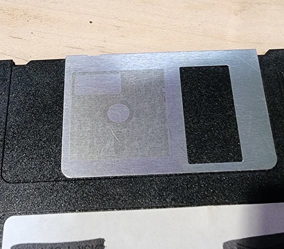 The metal shutter of a 3.5" floppy disk, onto which is etched a faint image of a 5.25" floppy disk 