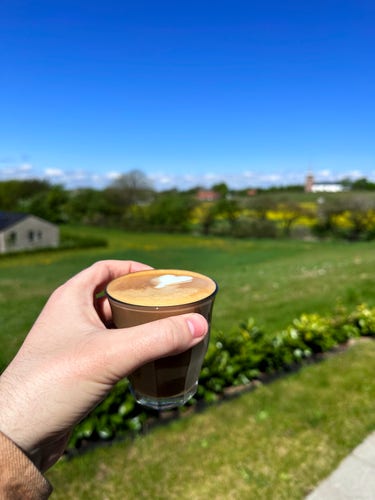 A hand holding a tiny espresso based drink with milk and foam. The photo is taken outside with a blue sky, green fields, and a church in the background.