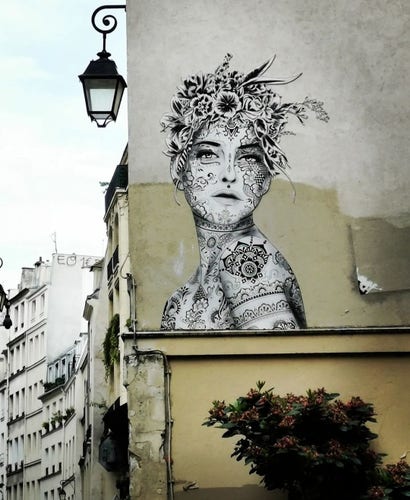 Streetartwall. A beautiful black and white mural of a young woman has been painted on part of the narrow beige and white exterior wall of an old five-storey building. The mural is on the second floor and shows the head of a woman looking over her shoulder. Her headdress consists of countless flowers and her face is also painted as if with tattoos. The rest of the visible body is also adorned with beautiful floral patterns.  (In the photo, a bush with flowers can be seen under the mural, a black street lamp hangs from the house on the left and the tall houses can be seen from the narrow alley).