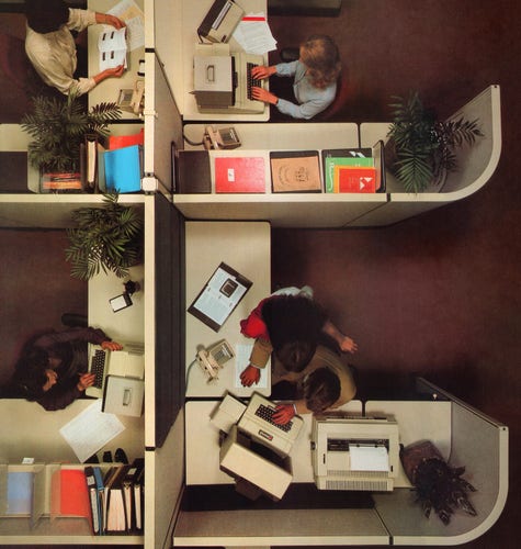 A photo of a few office cubicles, looking straight down from the ceiling. Poeple are wearing early 80s clothing, with Apple II computers, potplants, folders, and old phones about.