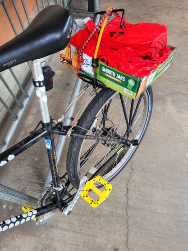 Mountain bike with a rack and yellow pedals with a ball canning box strapped on the rack with a big red shopping bag.