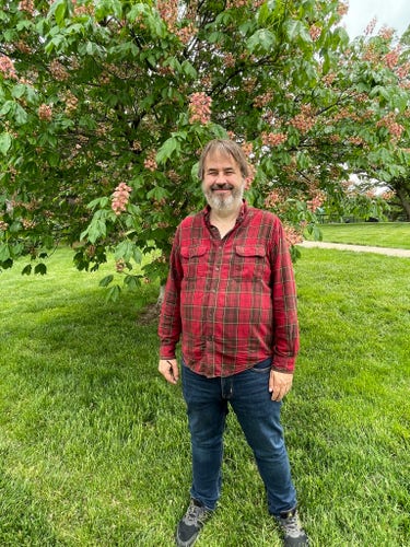 Chuck standing in front of a flowering tree. Red plaid shirt and jeans.