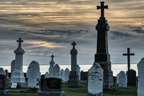 This is a photograph of a cemetery at either sunrise or sunset, with the sky displaying a palette of orange, gray, and blue hues. Numerous tombstones, marked with crosses, indicate it is a Christian burial site. The gravestones vary in design and show signs of age, suggesting a range of historical periods. A large, ornate Celtic cross stands prominently among them, suggesting its significance. The calm sea in the background meets the horizon under the soft sky, adding a tranquil and reflective ambiance to the scene.