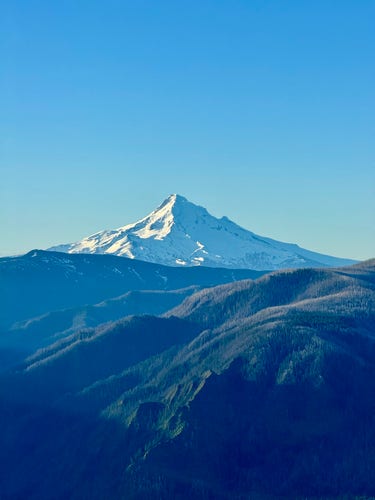 A beautiful snow-covered Mount Hood rises above the Central Cascades mountain range. The volcano has sunlight shining on the snow from the east (left) side. Blue sky on an amazing spring day. Vast green forests are seen before the peak. 