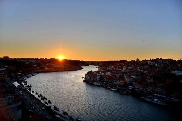 Zachód słońca nad rzeką z miastem i łodziami, z mostem widocznym w oddali.