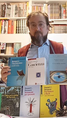 @andrsci con i libri di Dennett che ha in casa (sei, in due file da tre. Prima fila: "Rompere l'incantesimo", "Coscienza. Che cosa è", "L'io della mente"; seconda fila: "L'idea pericolosa di Darwin", "Strumenti per pensare", "Dai batteri a Bach").
AndrSci ha una camicia celeste e un gilet rosso. Sullo sfondo, una libreria Ikea piena di libri.