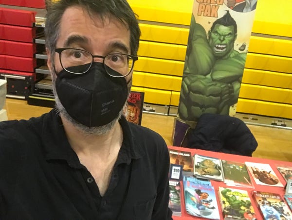 Greg Pak in a mask standing before his comics-laden table at the FanFaire NYC comic con.