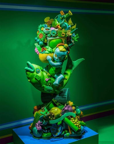 A colorful sculpture by British artist Gary Card featuring a small child riding a large fish. The sculpture is adorned with everyday objects and abstract shapes.