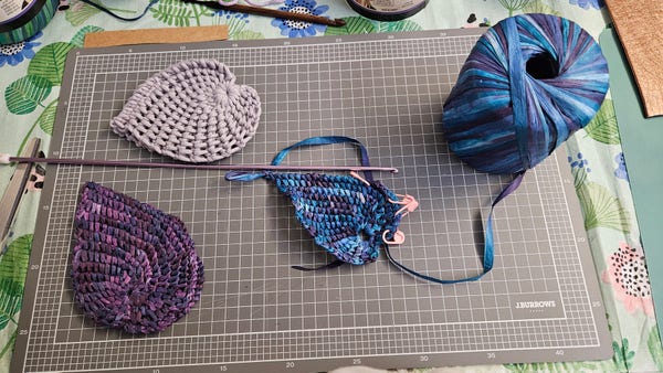 A grey cutting board with a white grid. On it sits three sets of crocheted curved leaves, one pale purple, one dark purple shades, and one shades of purple and blue. The third one  is still in progress with stitch markers. Beside it is a roll of raffia paper yarn in shades of blue and purple. Above it lies a long lavender crochet hook.
