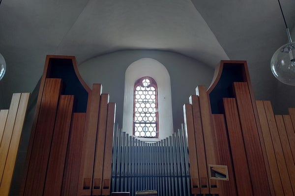Wir blicken auf ein Kirchenfenster, durch das helles Licht scheint. Im Vordergrund Orgelpfeifen aus Metall und aus Holz. Die Orgel steht genau mittig vor dem Fenster und die nach links und rechts größer werdenden Pfeifen scheinen es einzurahmen.