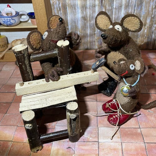Photo of Minimus, Minima and Silvius, the Latin mice, in a woodwork lesson. Minima has balanced a plank with peg holes on the kitchen chair. Minimus his helping Silvius to knock in a wooden peg with a hammer that is rather too big for his little paws