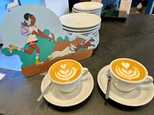 Two cappuccinos in white mugs at the counter in Ritual coffee. There is a sign with art of a woman using rollerskates and being pulled by a three leashed dogs as she listens to music on her headphones. 