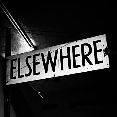 Black and white photo of a dark space with a sign that says "elsewhere" in all caps