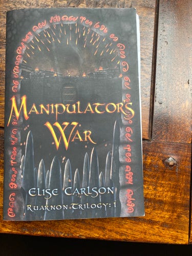 A book titled "Manipulator's War" by Elise Carlson, part of the RuarNon Trilogy, is lying on a wooden surface. The cover features stylized text and a dark fantasy-themed illustration with a silhouette of a castle. 