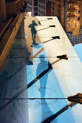 A reflection within a glass awning of two people walking along a sidewalk, buildings in the background flipped upside down extend downward into the sky.

The awning reflecting the sidewalk extends from the bottom of the frame forward, shrinking in perspective, like a road, ending well below the top edge. Along the awning, seams of the large panes of glass that make up the awning, each seam is met with a triangular mount of a support beam.

The person in the front is wearing headphones and a large puffy jacket. Their shadow extends behind them.

The second person behind them, larger because of the perspective, has short hair, a jacket, pants, white shoes. They shadow extends behind them beyond the bottom edge.

The sky is blue behind the second person, the buildings and sidewalk are illuminated with golden sunlight.