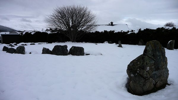 A standing stone in the right foreground, with a circular kerb of stones in the centre. The stones stand in deep snow today. Behind there are domestic hedges and rooftops. It's very grey and overcast, and all of the colour is gone.