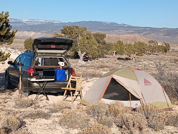 A color photo showing a car facing toward the left with it's back open. There's a table and a wood chair. There's items on the table, a book  camera, laptop, stove and pan. And a small tent is in the lower right of the frame.