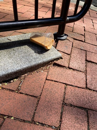 SNapshot of a brick sidewalk and metal gate or bike rack affixed to the ground. Underneath it is a clear cup containing milky coffee, laid on its side, not spilling because of its clear lid