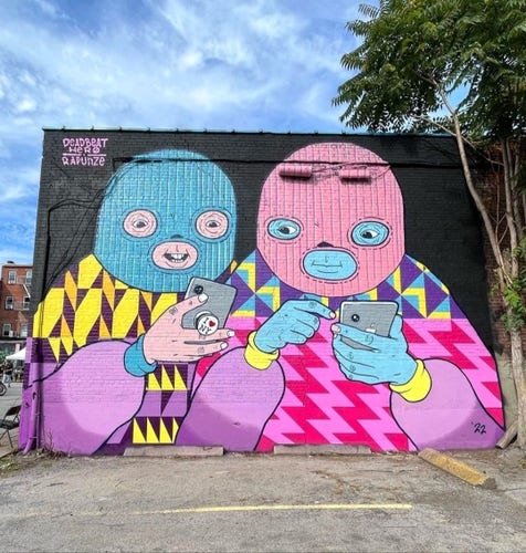 Streetartwall. The mural with two colorful masked figures with smartphones in their hands was sprayed/painted on the exterior wall of a one-story red brick building. The background of the colorful comic mural is painted black. Both figures are wearing woolly hats with three slits in the face and have large round eyes and mouths. The colors of the woolly hats are turquoise and pink, their clothes have zigzag and diamond patterns and are bright pink, yellow and purple. They look at the smartphones in their hands with interest. (The photo shows a parking lot in front of the mural).
