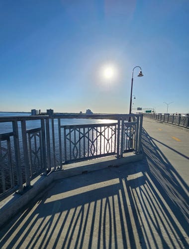 From an observation deck high above a vast river, the bright sun is low in a brilliant clear blue sky, casting a bright reflection across the water below and large, crawling shadows from the bridge railings onto the pavement and walkway.