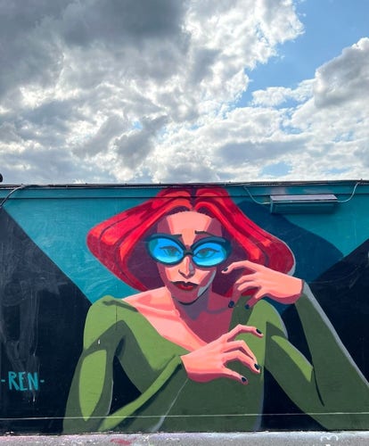 Streetartwall. A mural with a red-haired woman in comic style was sprayed on a long street wall. The background is geometrically divided into green and turquoise. The somewhat strange-looking woman has short red hair that is styled voluminously on both sides. She is wearing oversized blue glasses and a green top. One eyebrow is raised and her red mouth is pursed, giving her a slightly annoyed look. She gestures with both hands in the air as if to curse someone.(Above the mural wall is a blue cloudy sky)