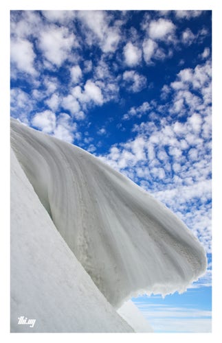 A large snowdrift has formed a large, thick, and overhanging frozen wave — almost Hokusai-like — seen from slightly below, and against a deep blue sky with streaks of little fluffy clouds. The lack of any other features gives the scene an almost abstract character.