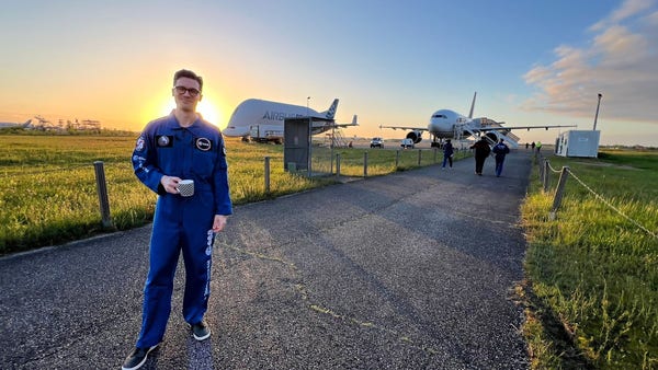 Actor and producer Steven Strait standing on a tarmac path in a blue ESA flight suit, with the sun behind him on the left and the Novespace Airbus 310 used to conduct zero-g parabolic arc flights. Steven is holding a cup of coffee, as is obviously needed.