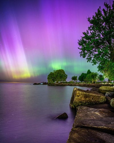 The aurora borealis over the Cuyahoga River in Cleveland. There are picturesque rocks and large stones, as well as leafy trees in the foreground. The aurora has lots of pink, purple, green, and even yellow.