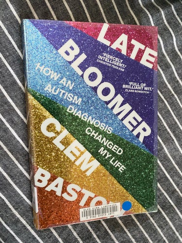 A book titled "Late Bloomer: How an Autism Diagnosis Changed My Life" by Clem Bastow, lying on a striped fabric. The cover has a glittery, rainbow design.