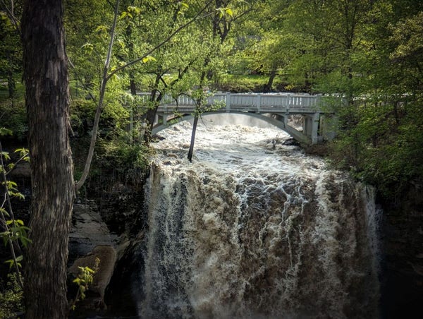 Photo of a raging waterfall flowing beneath a white concrete pedestrian bridge in the midst of a green forested park in spring.