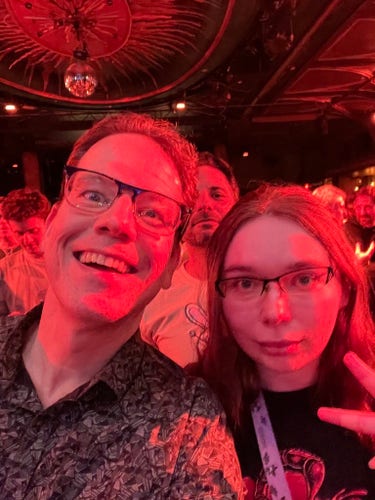 Your humble correspondent and a friend taking a goofy concert-floor selfie. An unknown and actually rather photogenic man is behind us, his face perfectly centered in-frame, looking directly into the camera, and smiling calmly.