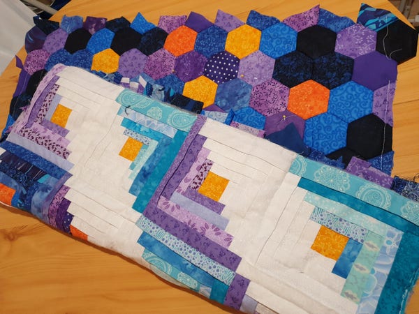 Photograph of a work in progress patchwork quilt, folded over asymmetrically to show the front with Manx log cabin in white, blues and purples with orange centre squares. The back, visible at the top, is English paper piecing with three inch hexagons in deeper blues, purples and with some orange and yellow. The edge is uneven with partial hexagons to make the back the same size as the front. 