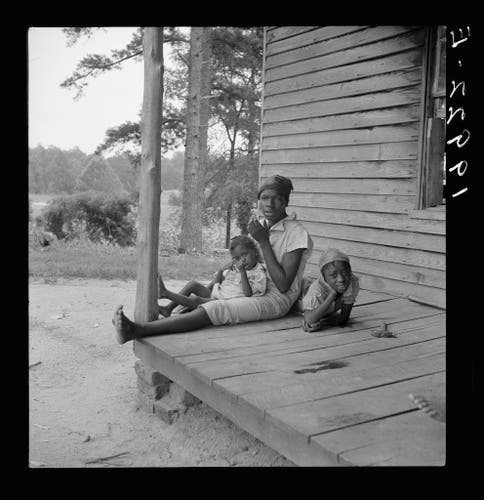  The image depicts a scene with three individuals, possibly a family, sitting on a porch. They appear to be outdoors in what might be a rural or semi-rural setting, given the structure of the house and the presence of trees. The person on the left is sitting down, seemingly engaged in an activity like eating or using a tool, while the two younger individuals are seated to their right, looking towards the camera with expressions that might be interpreted as curiosity or mild amusement.

The photo has been taken from a perspective slightly above eye level, which provides a clear view of the subjects and their immediate surroundings without any obstructions in the frame. The lighting suggests it could be either dawn or dusk, as the sunlight is soft but still brightly illuminating the scene.

The image has a vintage quality to it, with faded colors and a texture that might indicate it's an old photograph. There are no visible texts on the image itself to provide additional context or information about the subjects or location.