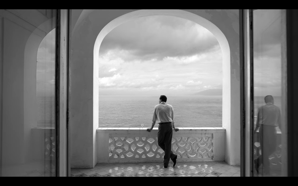 The character Tom Ripley stands, leaning against a balcony under a large arch, as he looks out to sea and a dramatic cloudy sky.