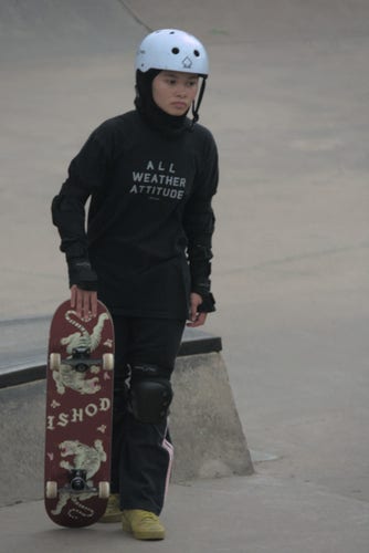 A young woman wearing a white skate helmet and all black clothing, looks serious as she steps forward holding her skateboard in her hand next to her, ready to drop it and roll off. The bottom of the skateboard is decorated with two stylish images of tigers, butterflies and the text ISHOD. 

Her shirt says "ALL WEATHER ATTITUDE" and her face shows it.