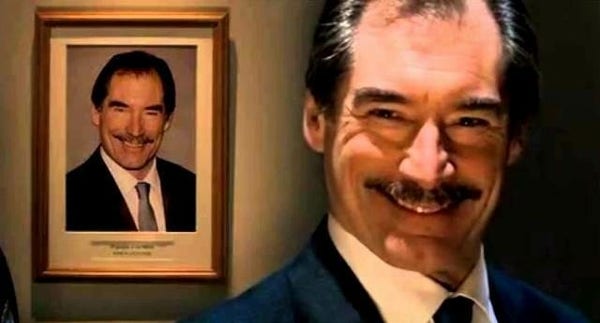 Timothy Dalton in "Hot Fuzz" as the supermarket manager. He's wearing a white shirt, jacket and tie, with a creepy insincere smile, and is standing front of a framed photo which has exactly the same clothing and pose.