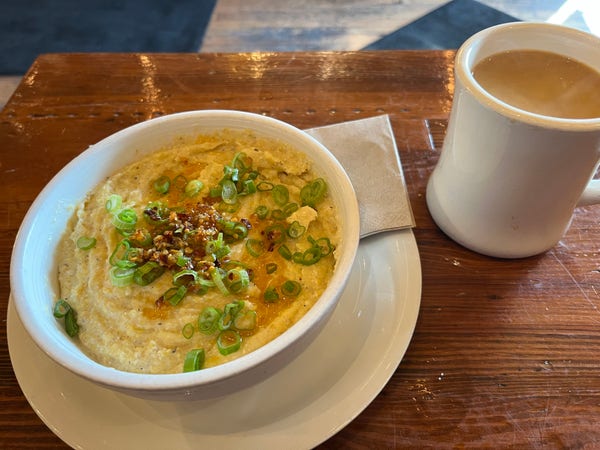 A bowl of grits topped with green onions and seasoning, beside a mug of coffee on a wooden table.