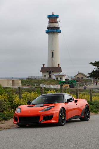 An orange exotic sports coupe in front of a lighthouse under a gray sky.