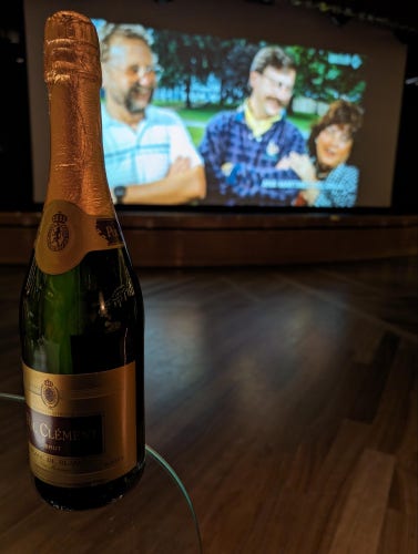 A bottle of champagne on a table in the foreground with a projector screen showing film footage presently with two men and a woman discernible even though they're out of focus.