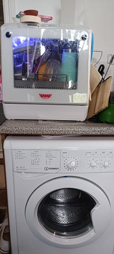 A washing machine machine in a tiny kitchen, where in a bigger house you might expect a dishwasher. on top, there's a tiny dishwasher, maybe a third or a quarter of the size of a regular dishwasher. it has a glass door, so you can see a bunch of dishes inside its blue lit interior. the dishwasher is decorated with two googly eyes no top, and a sticker of a mouth on the bottom 