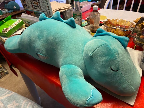 A large teal plush dinosaur toy on a red table with various items in the background including a bottle of soy sauce, a glass bowl, and some papers.