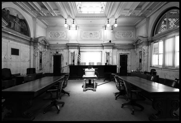 A monochrome, high contrast view of an empty, marble-clad, Greek revival, late 1800s federal courtroom.

Stark bright light from a skylight falls on a lectern and a large poduim desk at the head of the room. Behind the desk, three high-backed leather chairs and an American flag. 

Looming in the darkness, two long conference table on either side of the room lead the eye forward to the podium where the judges sit.