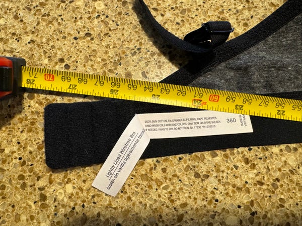 A photograph of the end of a bra’s band with the label showing a size of 36D. this bra should be slightly less than 36 inches (91.5cm) long so that there is a little bit of elastic grab when on a person with a 36 inch chest. There is a tape measure, laying on it showing that the bra is 27 1/2 inches, or 70cm long. That is beyond the end of the clips you have to use to secure it.